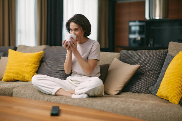 Modern girl drinking coffee or tea holding mug sitting on comfortable sofa, watching TV series, movie, resting at home Modern girl homeowner or tenant drinking coffee or tea holding mug sitting on comfortable sofa in living room, watching TV series, movie, resting at home. High quality photo Video Equipment Rental stock pictures, royalty-free photos & images
