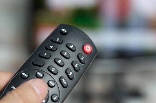 Changing TV program: close up of human hand holding television remote control on blured background with TV screen