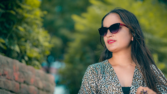An Asian/Indian beautiful young woman relaxes and enjoys fresh air and a tranquil scene on a sunny day in her leisure. She wears dark sunglasses and a spotted top.