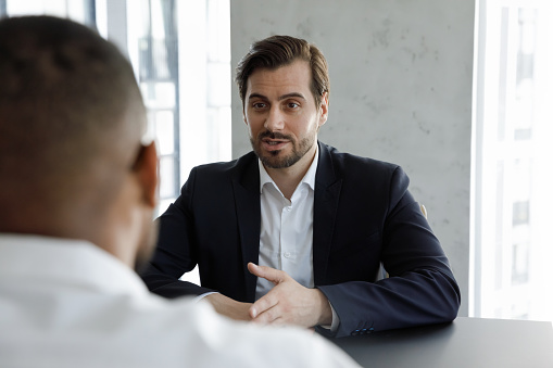 Concentrated young Caucasian man in formal wear involved in interview conversation with professional African American hr manager, discussing experience or job offer at meeting, headhunting concept