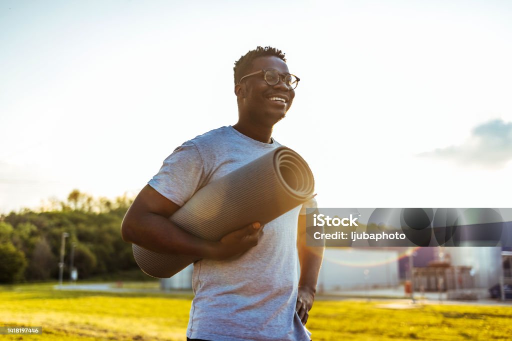 African american man holding yoga mat smiling on sunny day. Happy young smiling  man standing alone before a yoga session Yoga Stock Photo