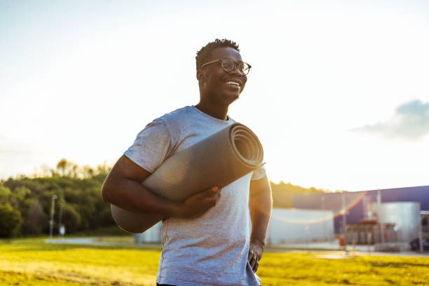 homem afro-americano segurando tapete de yoga sorrindo em dia ensolarado. - men exercising equipment relaxation exercise - fotografias e filmes do acervo