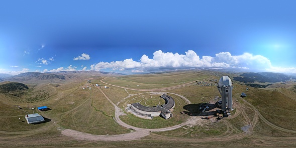 360 degree photo of the Assy-Turgen Observatory. A tall building with a dome and a telescope. Several houses and a tent camp. Big white clouds, mountains and hills covered with yellow-green grass