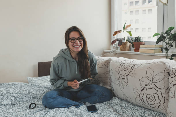 Young lady feeling happy about getting pregnant that she find from pregnancy test she is holding while sitting on bed. Woman with happiness facial expression future mother positive surprise from test. stock photo