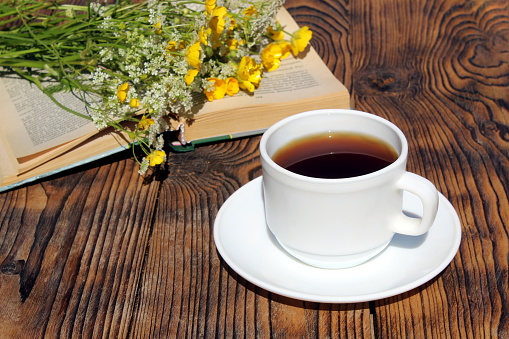 On a wooden table is a cup of tea with a book and a bouquet of wildflowers.