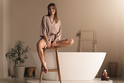 Cheerful young lady in elegant silk dressing gown sitting on wooden stool in stylish luxury bathroom against modern ceramic bathtub, ready to take bath, having beauty and spa procedures