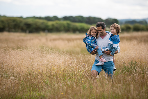 A medium close-up of a father carrying both of his kids in his arms as they enjoy a day out together in the Idyllic coastal town of Seahouses in the North East of England.