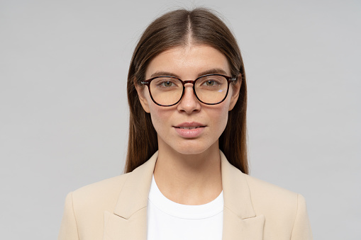 Headshot of intelligent businesswoman in glasses isolated on gray background with confident facial expression. Portrait of young smart successful CEO
