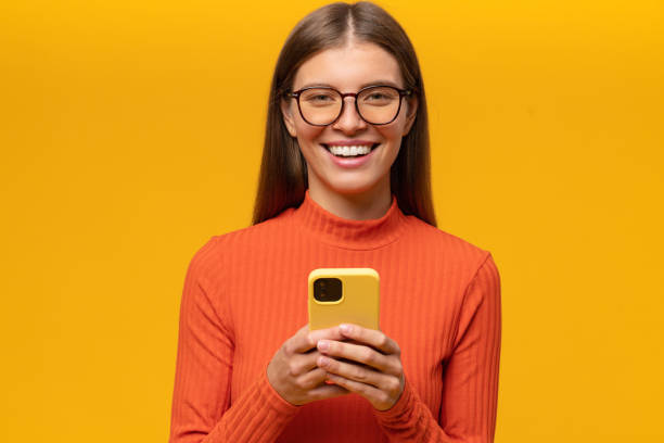 Woman in glasses posing on yellow background using phone laughing at memes in social media Pretty young woman in casual clothes and eyeglasses posing on yellow background using mobile phone, sending and receiving messages, reading comments in social media, laughing at memes i see you meme stock pictures, royalty-free photos & images