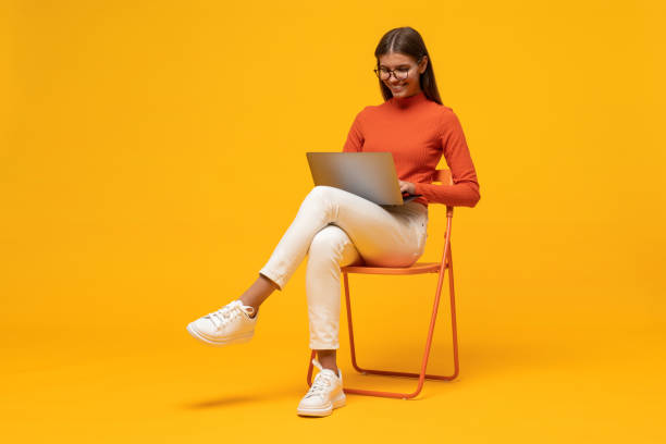 ritratto di studentessa che studia online seduta sulla sedia con il laptop sulle ginocchia su sfondo giallo - isolated objects foto e immagini stock