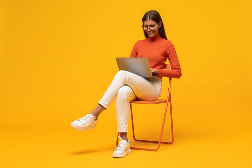 Side view of smiling attractive student girl studying online sitting on chair with laptop on knees on yellow background with copy space. Modern education and technology