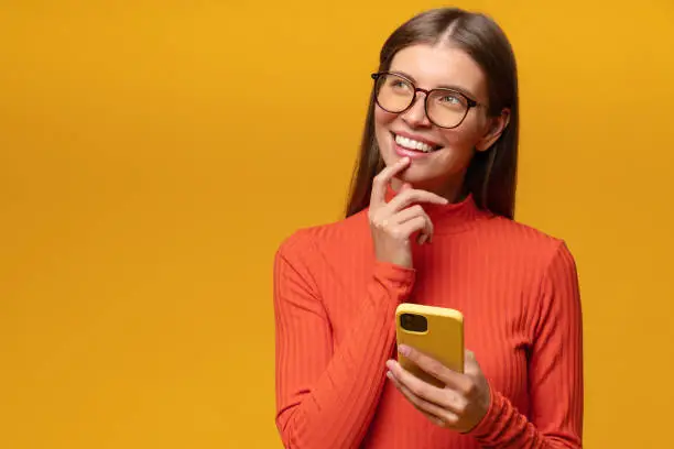 Photo of Portrait of thoughtful woman looking up aside writing message to guy in dating app holding phone