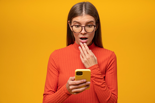 Studio image of shocked woman of 20s reading message with petrifying news, touching opened mouth, looking at screen with shocked face on yellow studio background