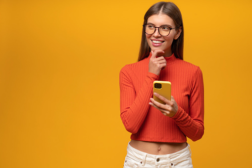 Pretty girl of 20s thinking while writing post for social media network holding smartphone in hands isolated on yellow background, looking aside with pensive face on copy space