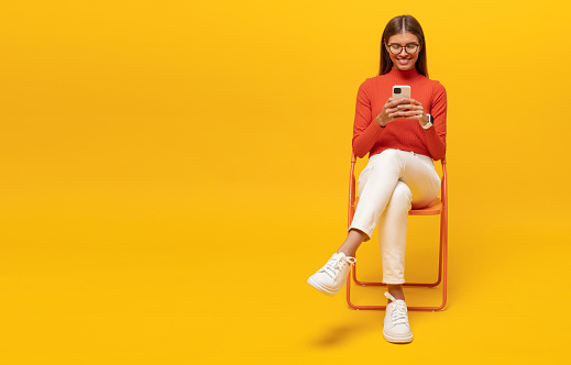 Full length horizontal portrait of millennial girl sitting at chair surfing mobile internet using wireless connection on yellow studio background with copy space