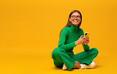 Dreamy student girl sitting on floor with phone isolated on yellow copy space background