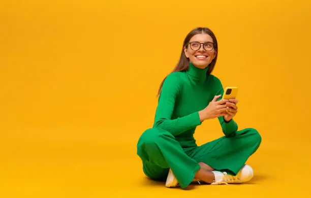 Photo of Dreamy student girl sitting on floor with phone isolated on yellow copy space background