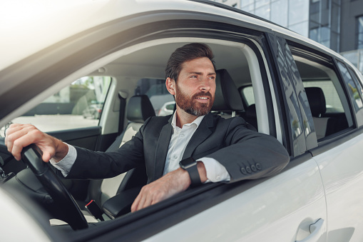 Handsome businessman in grey suit is riding behind steering wheel of car. Blurred background