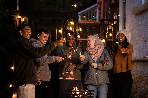 Young men and women are outside in a garden holding sparklers together around a fire. Friends celebrate at a social gathering lauging and smiling wearing warm clothing.