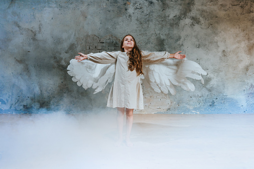 Little girl in ethnic dress wearing angel wings standing in white smoke raising her hands and looking up.