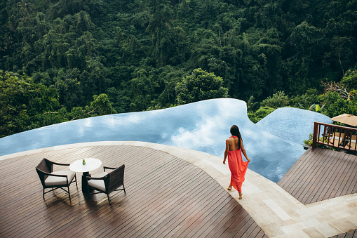 Aerial shot of young female model in sundress walking by the swimming pool. Woman at luxury resort poolside.