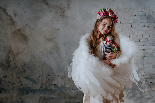 Little girl with angel wings and flowers wreath holding traditional handmade Ukrainian fabric doll. Selective focus. Loft background with blank space for text.