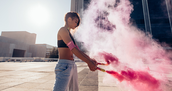 Sporty young woman holding smoke sticks outdoors in city. Sportswoman holding a smoke grenades over urban city background.