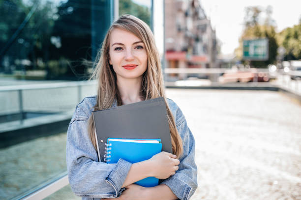 a young girl employee is standing in the city on the street on a sunny day, smiling and holding folders and notebooks in her hands against the background of a glass business center - estudante universitária imagens e fotografias de stock