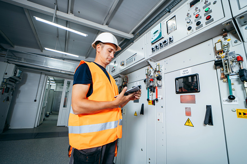Adult electrician builder engineer testing and screwing equipment in fuse box and repairing of modern electricity power station using data from the tablet.. Automatic control cabinet