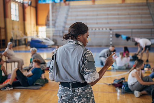 Diverse group of people, soldiers on humanitarian aid to civilians in school gymnasium, after natural disaster happened in city. Female soldier counting civilians.