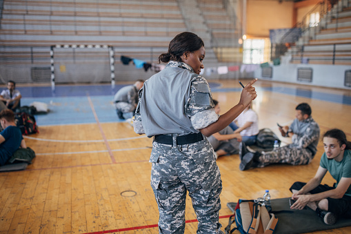 Diverse group of people, soldiers on humanitarian aid to civilians in school gymnasium, after natural disaster happened in city. Female soldier counting civilians.