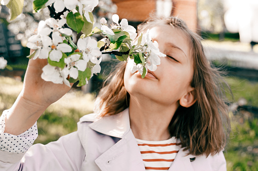 Little girl is walking in spring garder with apple trees and white flowers. Romantic stylish spring kid in coat. Blooming in sunny park. Beautiful nature photoshoot. Spring bloom enjoy, relax.