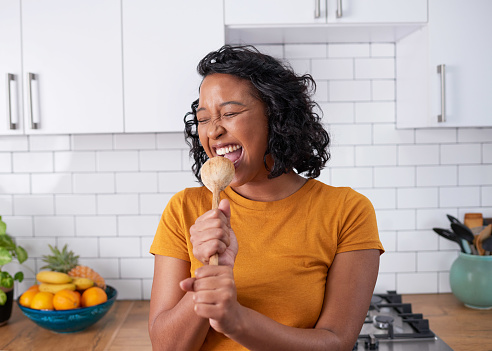 A young multi-ethnic woman sings into wooden spoon in modern kitchen. High quality photo