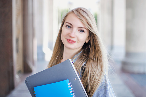 Female student holding a notebook, folders and smiling against the background of university, college