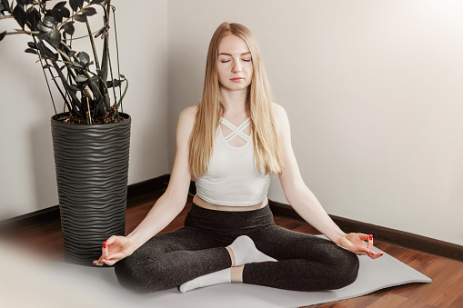 Young Caucasian blond woman sitting in lotus position on yoga mat with closed eyes and meditating at home. Healthy lifestyle during quarantine
