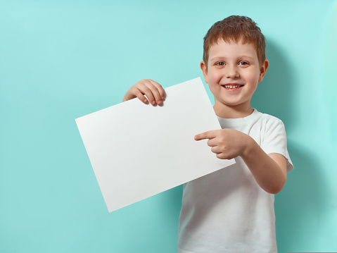 Four-year-old boy smiles and shows finger on blank white sheet. Happy child on blue background with copy space for message, mock up