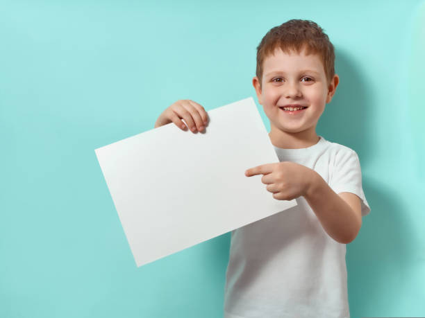 Little boy holds blank paper sheet, blue background stock photo