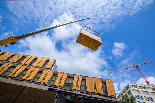 A crane with spreader frame lifting a building block of a temporary prefabricated sustainable timber modular office structure in a city environment.