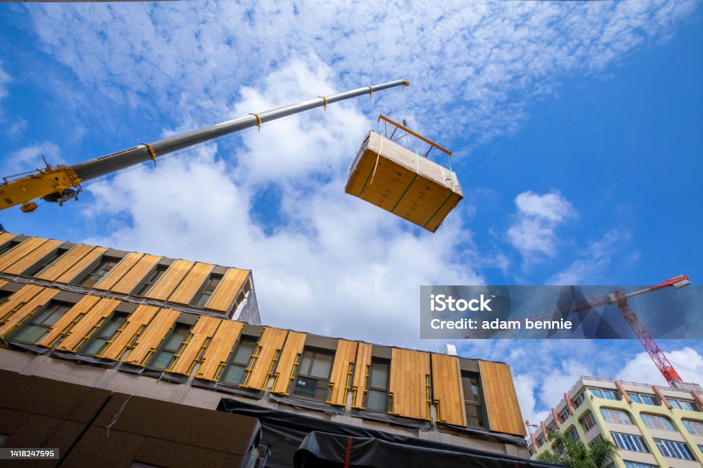 Pre-fabricated modular wood commercial building being assembled with crane A crane with spreader frame lifting a building block of a temporary prefabricated sustainable timber modular office structure in a city environment. Construction Industry Stock Photo