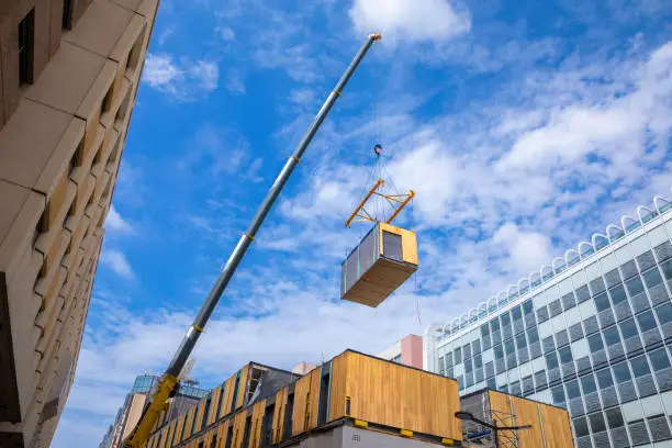 A crane with spreader frame lifting a building block of a temporary prefabricated sustainable wood modular office structure in a city environment.
