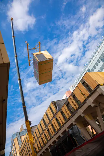 A crane with spreader frame lifting a building block of a temporary prefabricated sustainable timber modular office structure in a city environment.