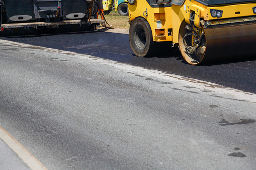 Close-up of Asphalt paving machine and road construction crew resurfacing suburban road with a new layer of asphalt. Workers leveling the asphalt with gravels. Steamroller in the foreground.