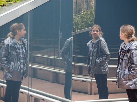 The girl is reflected on the mirror surface.  A teenage girl in a gray jacket on vacation looks at a wall with a mirror surface that reflects her.  A scene from real life