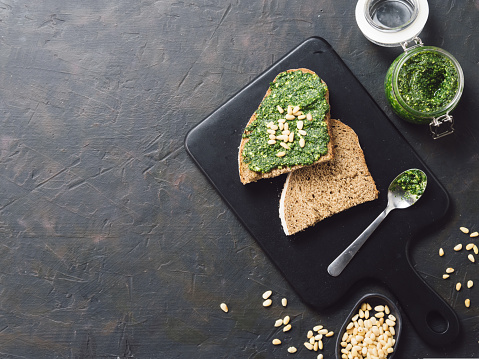 whole grain rye bread with fresh basil pesto sauce on black wooden cutting board over dark concrete background. Top view or flat-lay. Copy space.