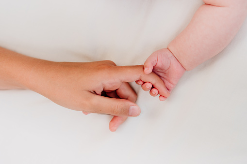 Newborn baby's hand holding adult's finger on white textile background with blank space for text. Top view, flat lay.