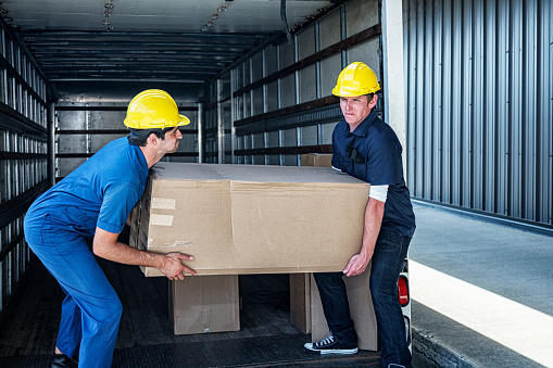Two young men - blue collar workers wearing work clothes and bright yellow protective headwear hardhat safety helmets - are working together; straining while carrying a very heavy cardboard box they are unloading from the back of an open truck trailer parked at a concrete loading dock. The man on the right is carrying the load with better technique - leveraging muscles in his whole body to help his straight arms. The technique and positioning of the man on the left is not ideal because he's carrying mostly with his arms rather than his whole body - potentially causing injury by putting undue strain on his arm, shoulder and back muscles.
