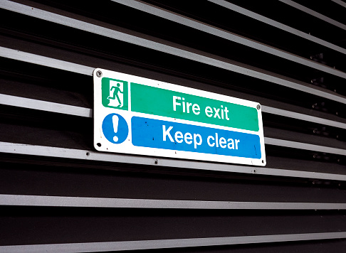 Green, blue and white sign on a louvered door reading ‘Fire Exit - Keep Clear’.