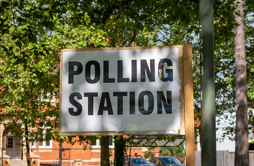 A Polling Station sign on a pole beneath trees outside a library on an election day in the UK.