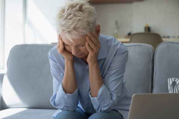 istock Upset older woman holding head with hands seated on sofa 1418260601