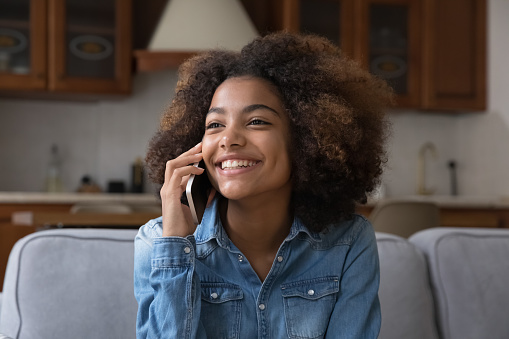 Beautiful curly-haired African teenage girl sits on couch talking on smartphone, holding mobile phone enjoy pleasant personal conversation, communicates remotely with boyfriend use modern tech concept